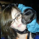 Monkey sits on the shoulder of a person while kissing her cheek.